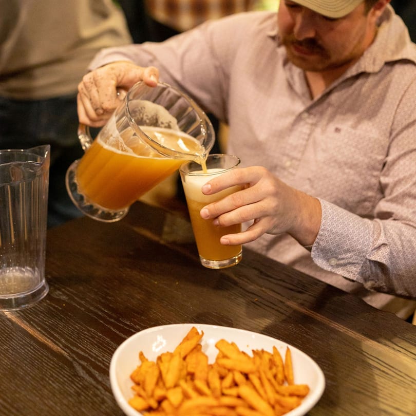 A person pours a pitcher of golden beer into a glass next to a bowl of fries.