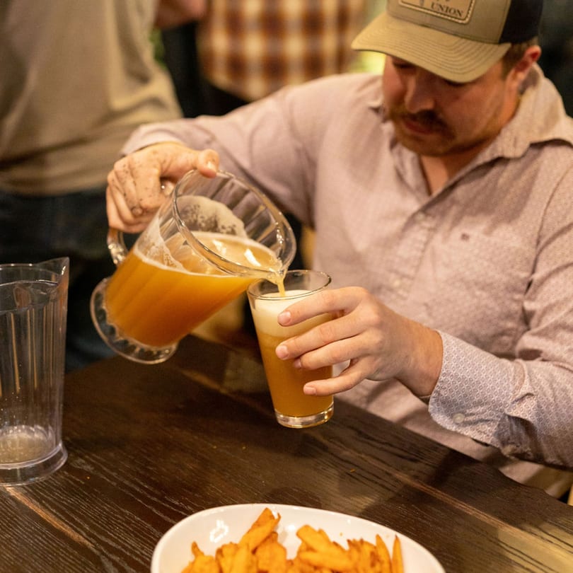 A man in a cap pours a pitcher of amber beer into a glass, with fries on the table.
