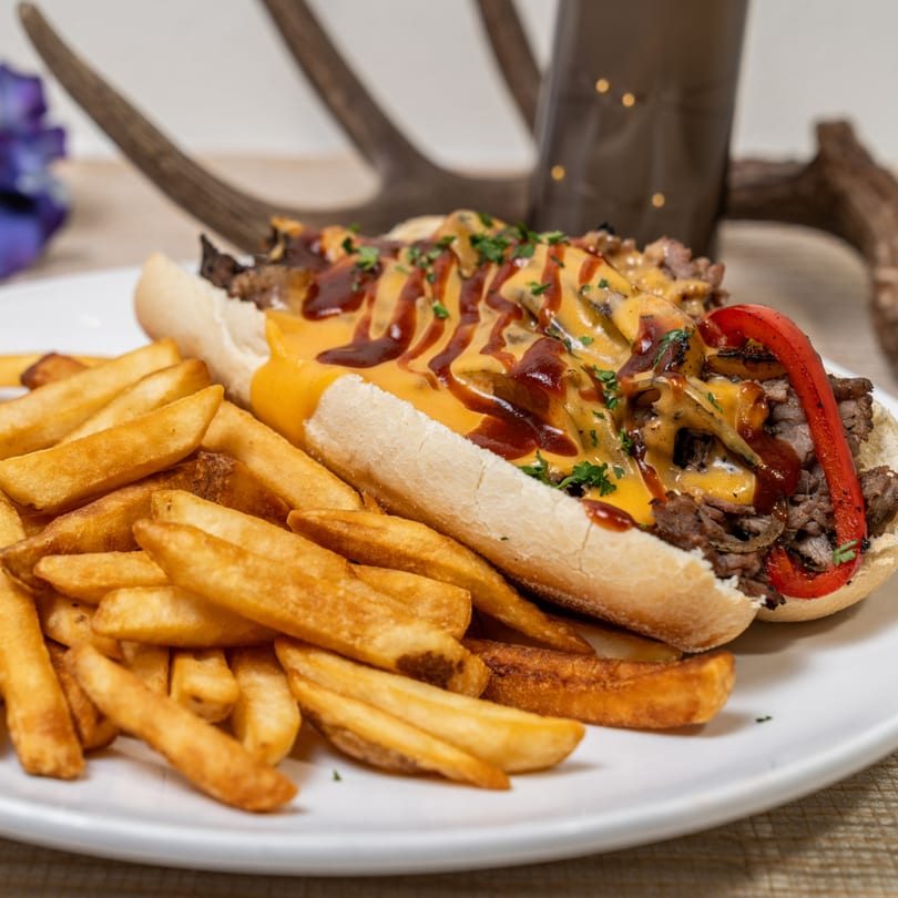 Cheesesteak sandwich with melted cheese, BBQ sauce, and red peppers, served with golden fries.