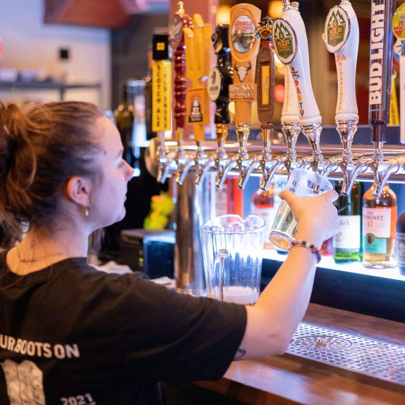 Bartender pouring a beer from a tap, with many beer taps and bottles in the background.