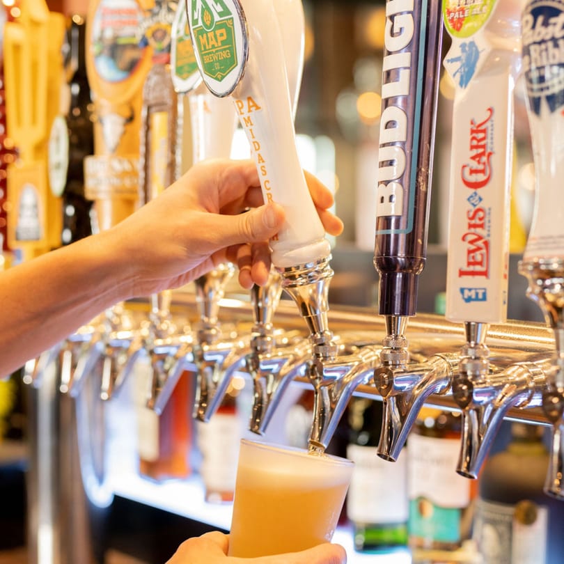 A hand pouring a light-colored beer from a tap into a glass at a bar.