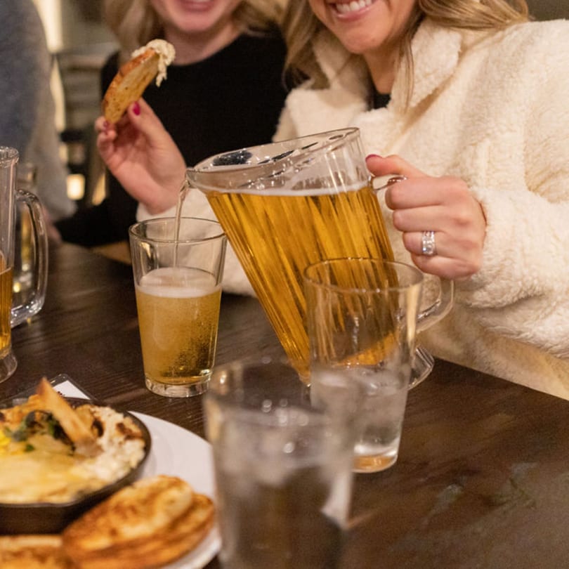 A person pouring beer from a pitcher into a glass at a table with food.