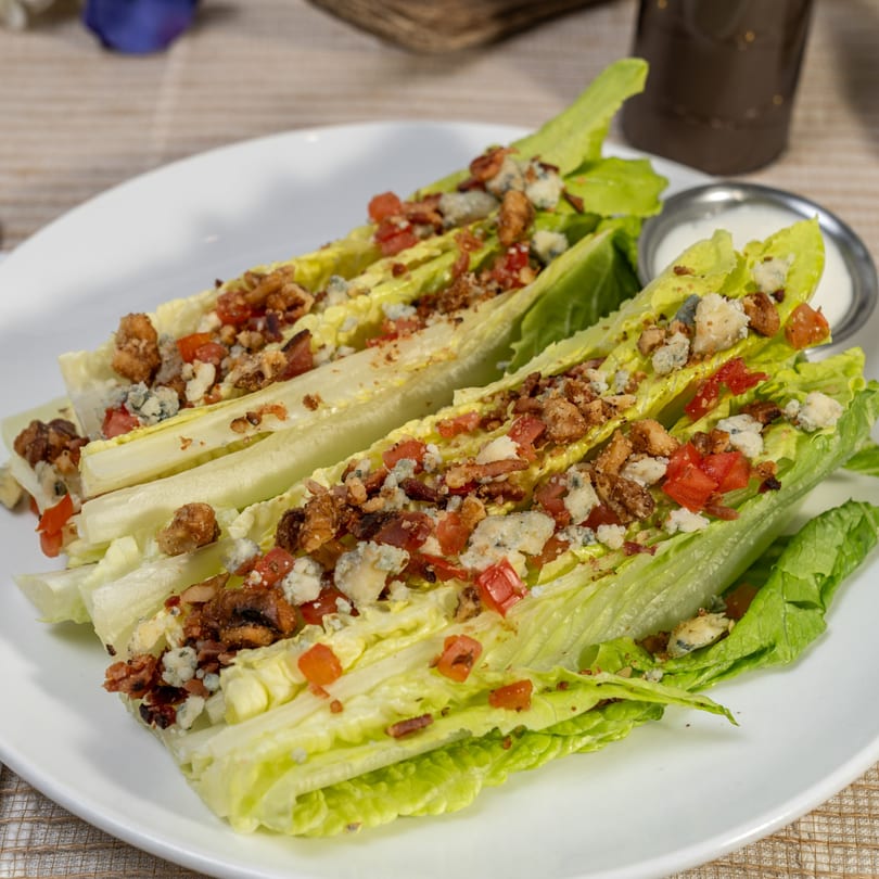 Two romaine lettuce wedges topped with bacon, blue cheese, and diced tomatoes, with a side of dressing.