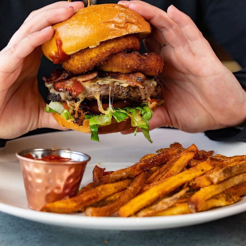 Hands holding a bacon cheeseburger with onion rings, served with sweet potato fries and ketchup.