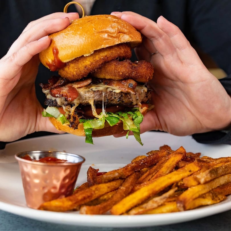 Hands holding a juicy burger with melted cheese, bacon, onion rings, and lettuce, served with fries.